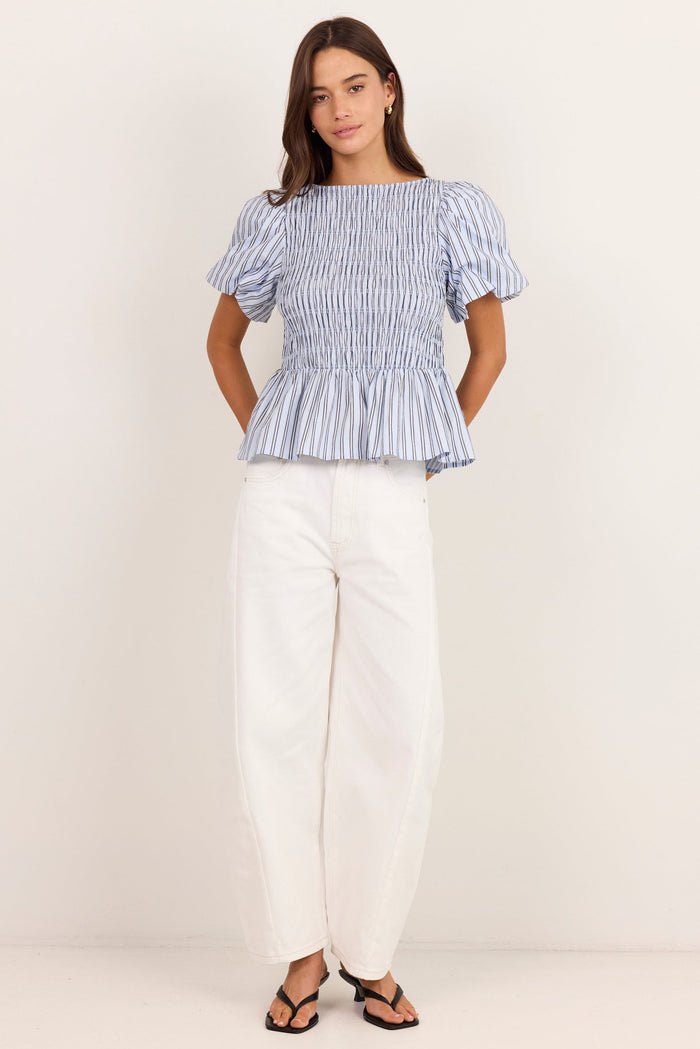 A woman modeling a blue and white vertically striped smocked peplum top with puff sleeves, paired with white wide-leg pants and black sandals, standing against a plain off-white studio background.