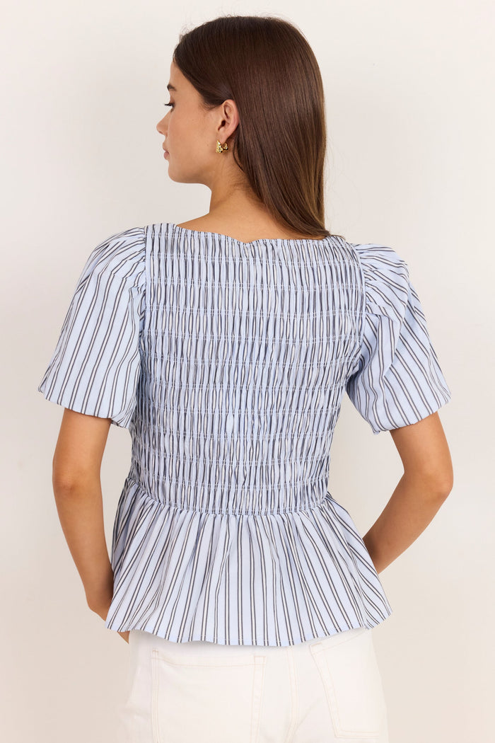 A woman shown from the back wearing a blue and white vertically striped smocked blouse with puff sleeves and a peplum hem, paired with white pants, against a plain off-white background.
col_pattern