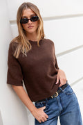 Woman wearing a brown sweater and blue jeans against a white wall.