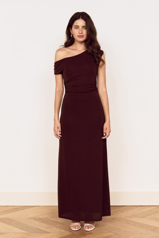 A woman modeling a floor-length off-the-shoulder dress in a deep burgundy or maroon shade, standing against a plain off-white wall on a light wood floor.