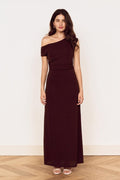 A woman modeling a floor-length off-the-shoulder dress in a deep burgundy or maroon shade, standing against a plain off-white wall on a light wood floor.