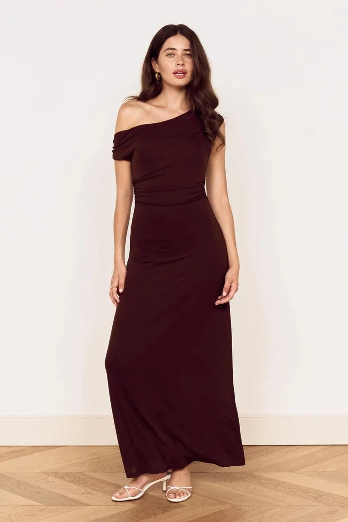 A woman modeling a dark brown off-the-shoulder maxi dress with a ruched waist, standing against a plain off-white wall on a light wood herringbone floor.