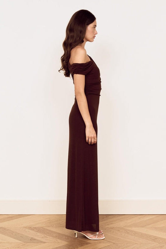 A woman stands in profile wearing a floor-length off-the-shoulder dark brown dress with a gathered waist, paired with clear high-heeled sandals, against a plain off-white wall and light wood herringbone floor.