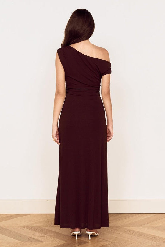 A woman viewed from the back wearing a dark burgundy one-shoulder maxi dress, standing on a light wood herringbone floor against a plain off-white wall.