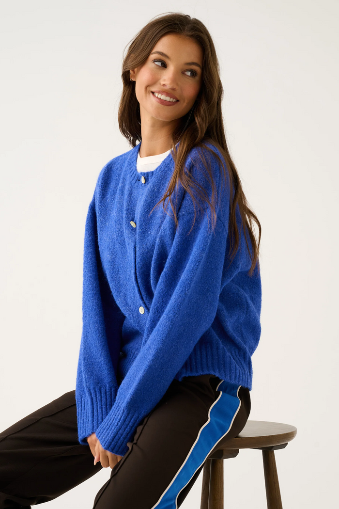 Woman wearing a blue cardigan sitting on a stool against a white background
