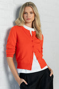 A blonde woman wearing a bright orange cardigan over a white top and black trousers, leaning against a white brick wall.