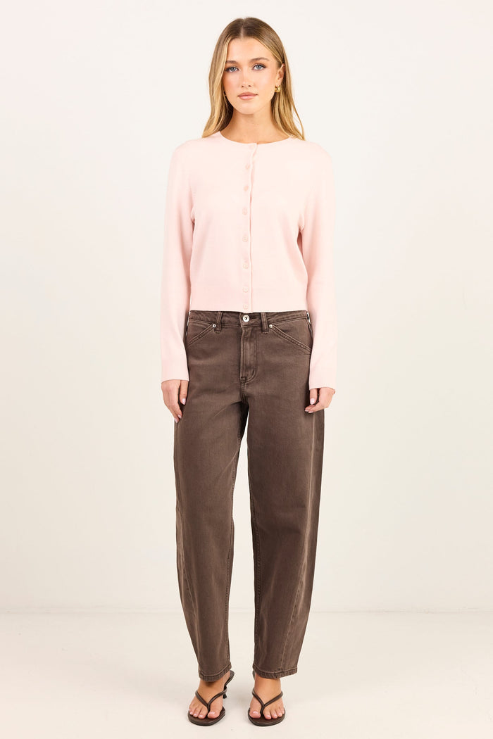 A woman stands against a plain light background wearing a pale pink long-sleeve button-front top, dark brown straight-leg jeans, and black sandals.
