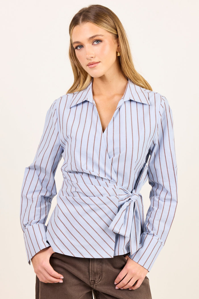 A woman modeling a light blue wrap-style button-up shirt with thin red vertical stripes and a tie waist, paired with brown pants, against a plain off-white studio background.