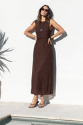 A woman wearing a sleeveless dark brown maxi dress and sunglasses stands with hands on hips in front of a white wall next to a pool.
