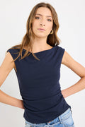 Woman wearing a fitted navy sleeveless top with boat neckline and side ruching, paired with light blue jeans.