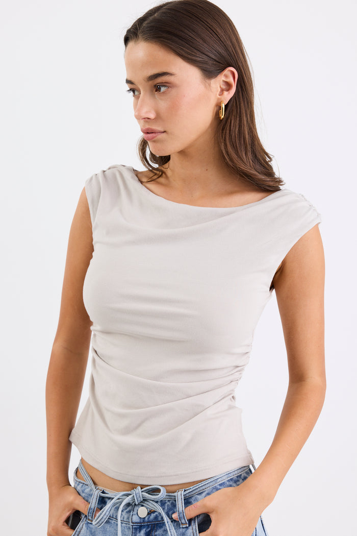 Woman wearing a stone-coloured sleeveless stretch top with a boat neckline, side ruching, and a fitted silhouette, paired with light blue jeans.