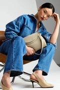 A model sits on a wooden chair wearing a blue denim jacket and matching wide-leg jeans, holding a beige shoulder bag, and wearing beige slingback heels against a studio background.
