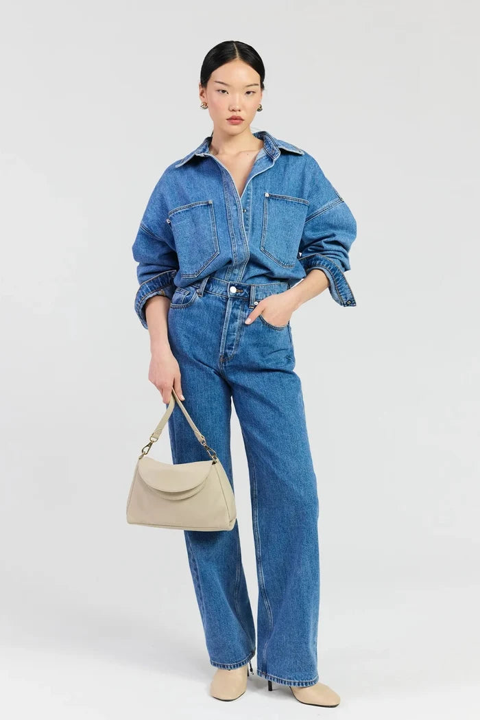 A model stands against a light gray studio background wearing a blue denim jumpsuit with wide legs, paired with beige pointed-toe heels and holding a beige handbag.