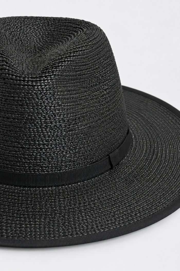 A close-up of a black woven straw fedora hat with a matching black ribbon band, shown against a plain light gray background.