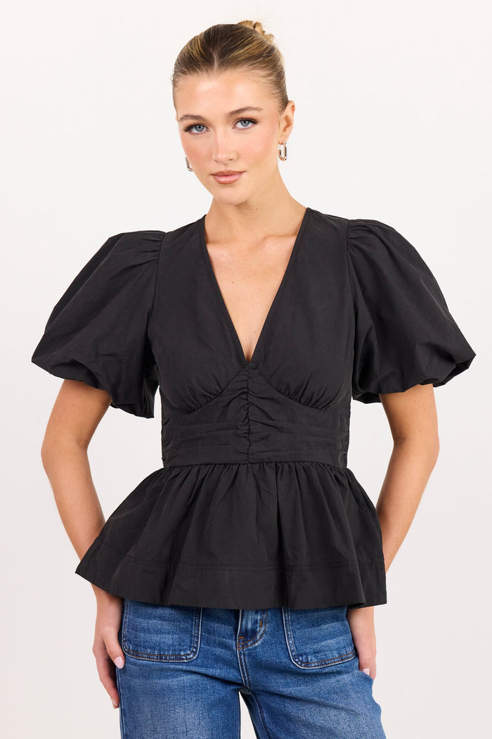 A woman modeling a black puff-sleeve peplum top with a deep V-neck and gathered waist, paired with blue jeans, against a plain light background.