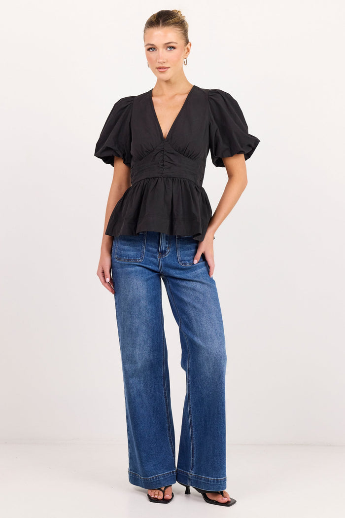 A woman modeling a black short-sleeve peplum top with puff sleeves and blue wide-leg jeans against a plain white studio background.