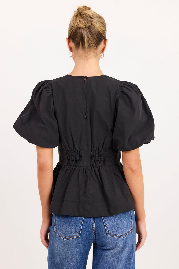 A woman viewed from the back wearing a black puff-sleeve peplum top with a smocked waist and blue jeans, against a plain white studio background.
col_pattern