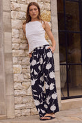 Woman wearing a white sleeveless top and navy floral pants leaning against a stone wall.