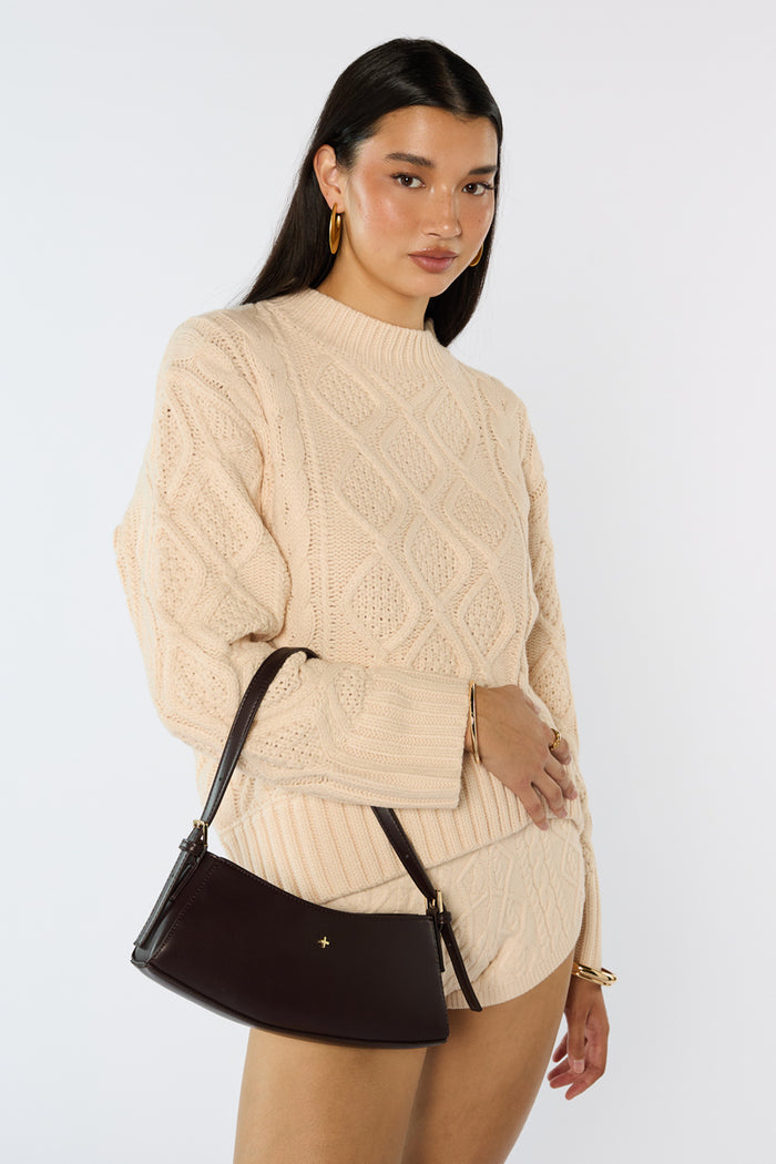 Woman wearing a beige cable knit sweater and holding a black handbag against a white background
