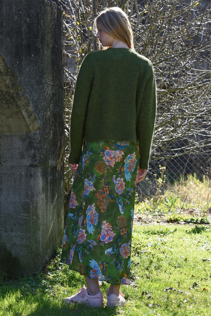 Person wearing a green cardigan and floral skirt standing outdoors near a stone wall.