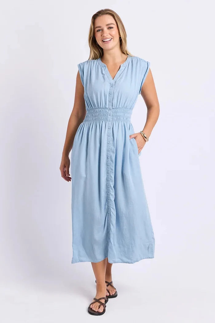 A woman modeling a light blue sleeveless midi dress with a smocked waist, buttoned front, and side pockets, photographed against a plain white studio background.