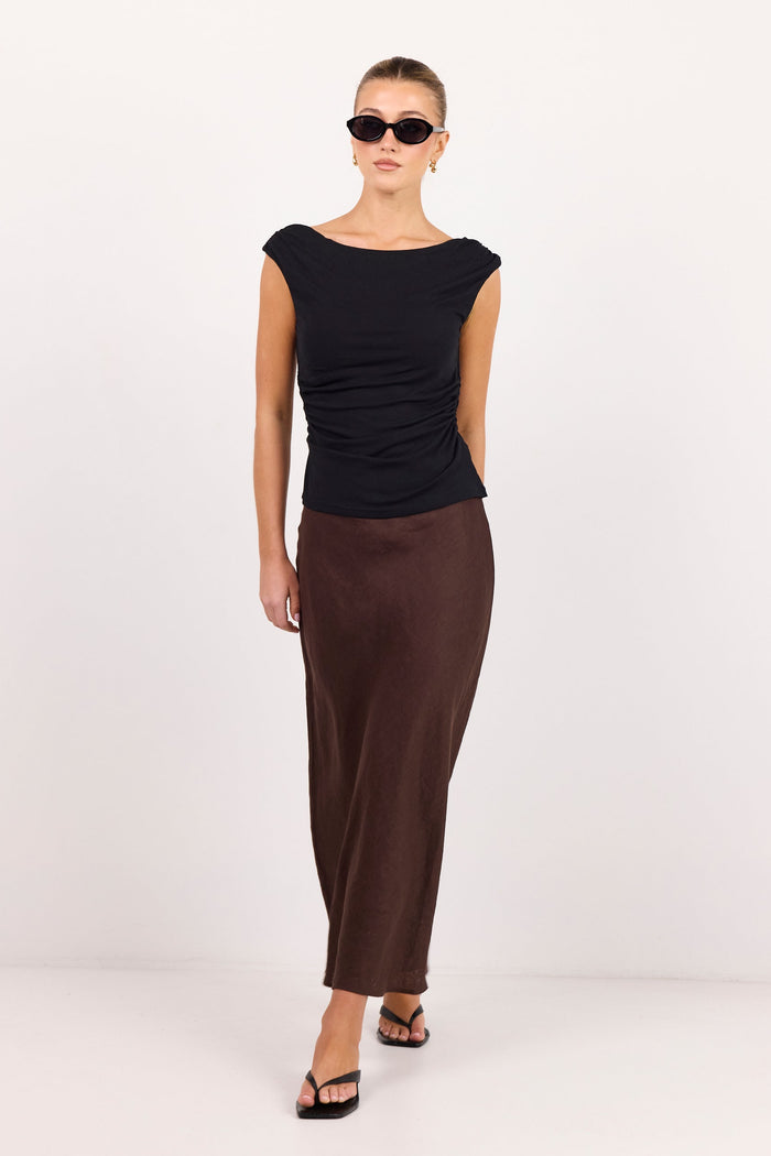 A model wearing a black cap-sleeve top and a dark brown satin midi skirt, styled with black sunglasses and black sandals, standing against a plain white studio background.