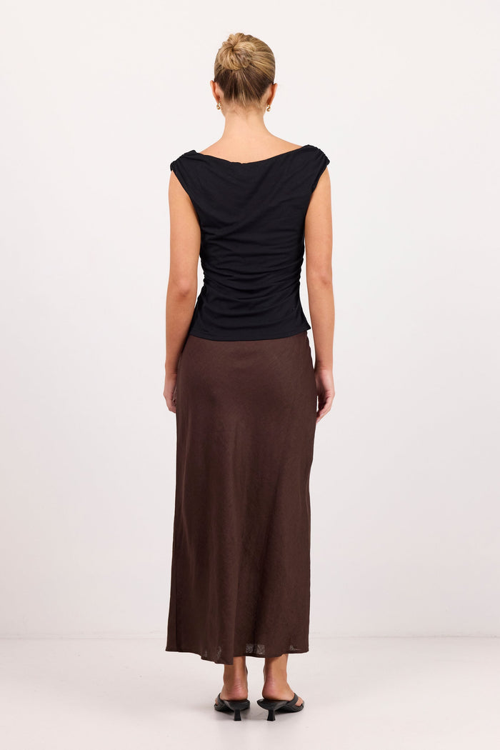 A woman viewed from the back wearing a black draped sleeveless top and a long brown skirt, standing against a plain light gray studio background.