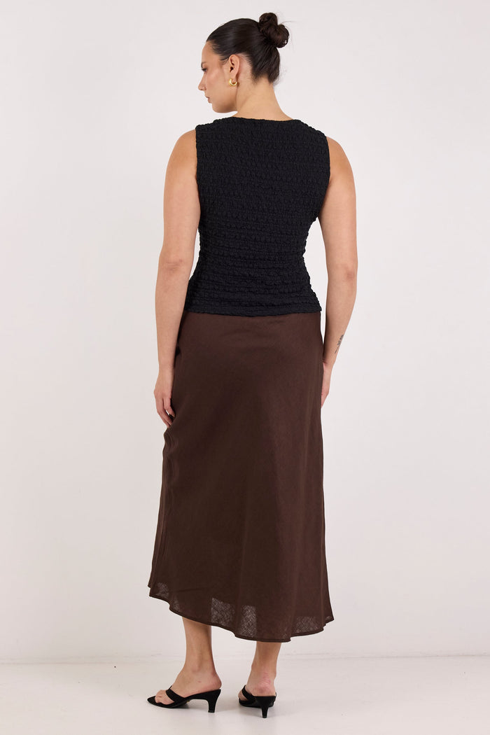 A woman is shown from the back wearing a sleeveless black textured top and a brown midi skirt, paired with black flat sandals, against a plain white studio background.