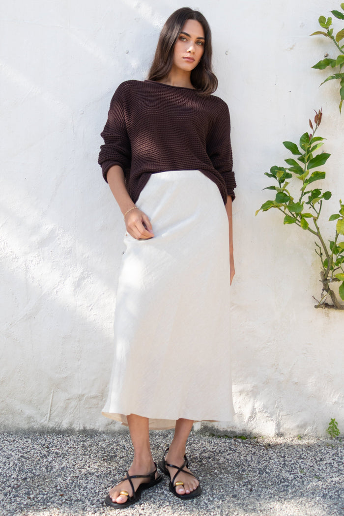 Woman wearing a brown top and white skirt standing against a white wall with a plant to the side.