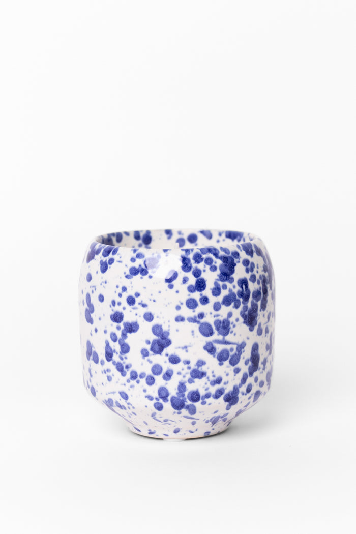 A small cylindrical ceramic cup with a white glaze covered in blue speckled spots, photographed on a clean white background.