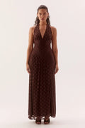 A woman modeling a dark brown halter-neck maxi dress with a subtle polka dot pattern, standing against a plain off-white studio background.