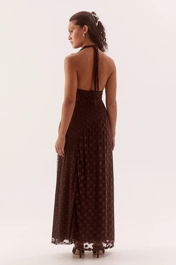 A woman modeling a dark brown halter-neck maxi dress with a subtle polka dot pattern, shown from the back against a plain white studio background.