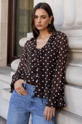 Woman in a polka dot blouse and jeans leaning against a building.