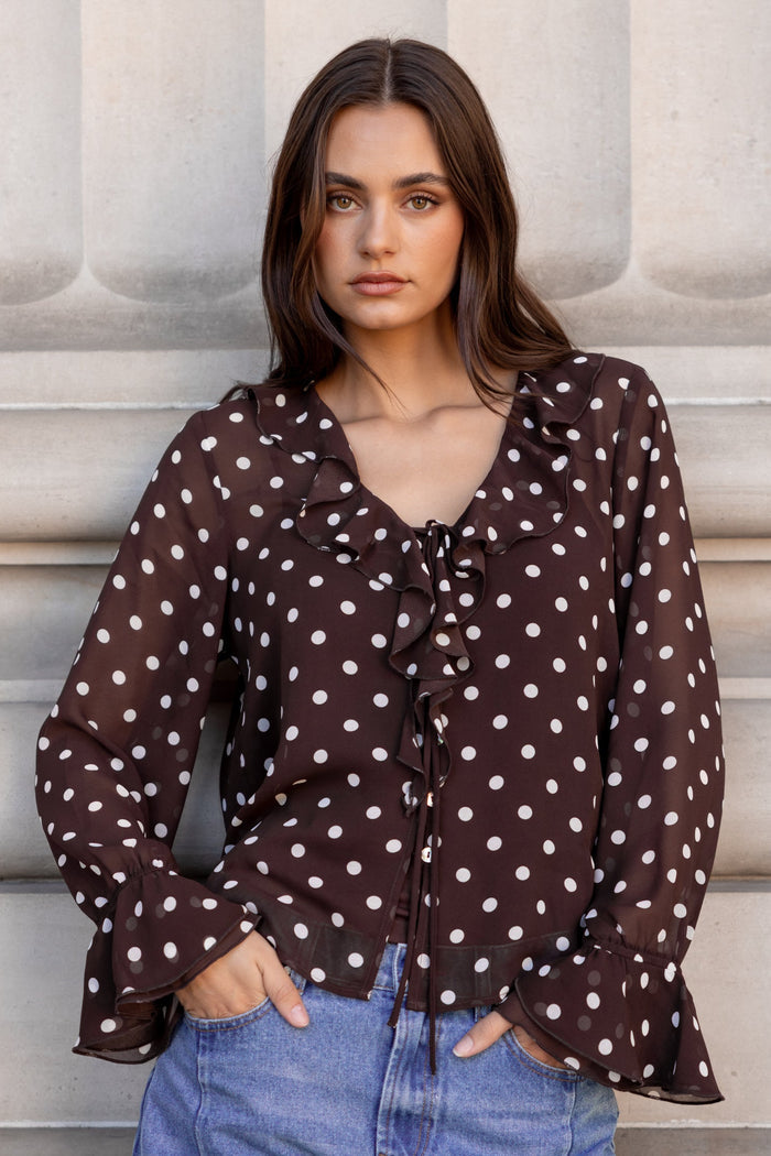 Woman wearing a brown polka dot blouse and blue jeans against a light-colored wall.