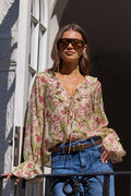 A woman wearing a beige floral long-sleeve blouse with tie-front detailing, blue jeans, and sunglasses, standing on a balcony with white architectural columns.