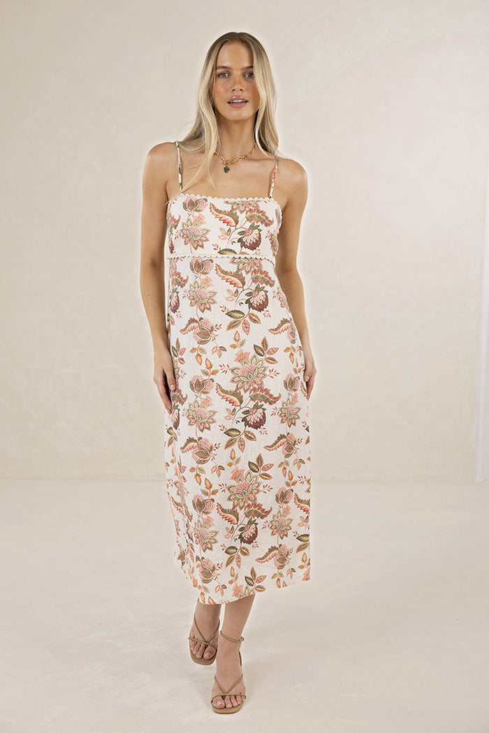 A woman modeling a sleeveless midi slip dress with thin straps and an allover floral and botanical print in muted pink, beige, and brown tones, set against a plain cream studio background.