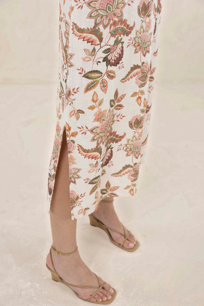 A close-up of a person wearing a long floral skirt with a side slit and tan strappy heeled sandals against a light neutral background.