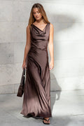 Woman in a brown evening gown holding a handbag against a light gray wall.