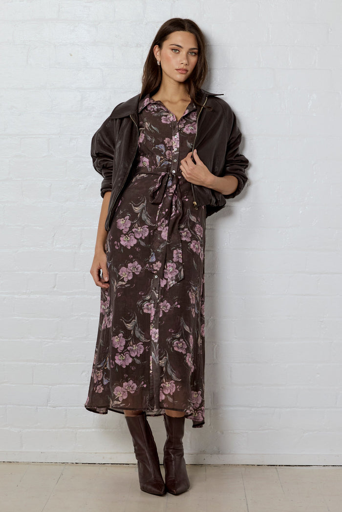 Woman wearing a brown floral dress with a brown leather jacket  against a white brick wall.