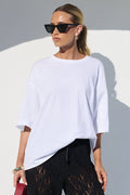 A woman modeling a white oversized short-sleeve top and black wide-leg lace pants, holding a red handbag, set against a minimalist white architectural background with strong sunlight and shadows.