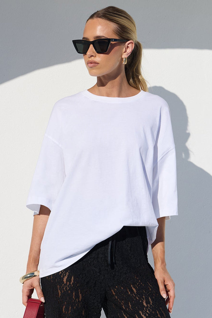 A woman modeling a white oversized short-sleeve top and black wide-leg lace pants, holding a red handbag, set against a minimalist white architectural background with strong sunlight and shadows.