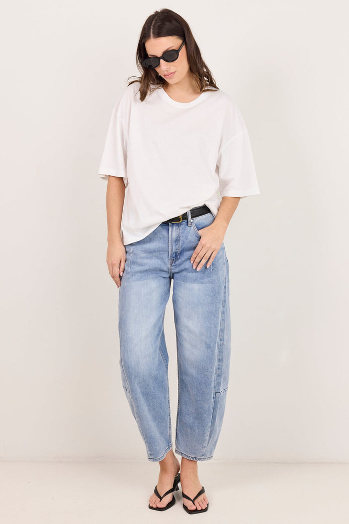 A woman modeling a white oversized short-sleeve t-shirt, light-wash wide-leg blue jeans, a black belt, and black sandals against a plain off-white studio background.