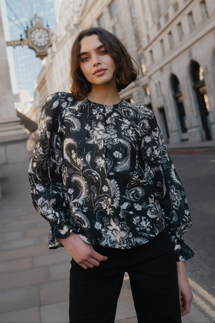 Woman wearing a black floral blouse standing in an urban setting with classical architecture.