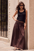 Woman in black tank top and brown wide-leg pants standing against a beige wall.