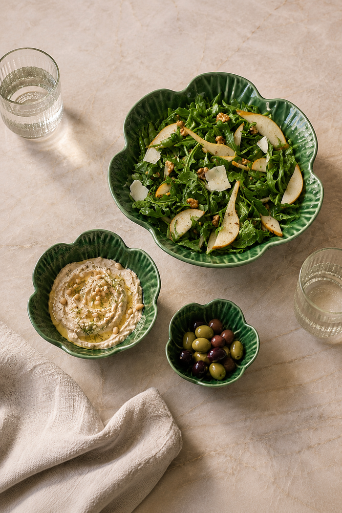 Three green scalloped ceramic bowls arranged on a light beige surface with a folded linen napkin, a glass of water, and a salad of greens with pears, hummus, and olives.