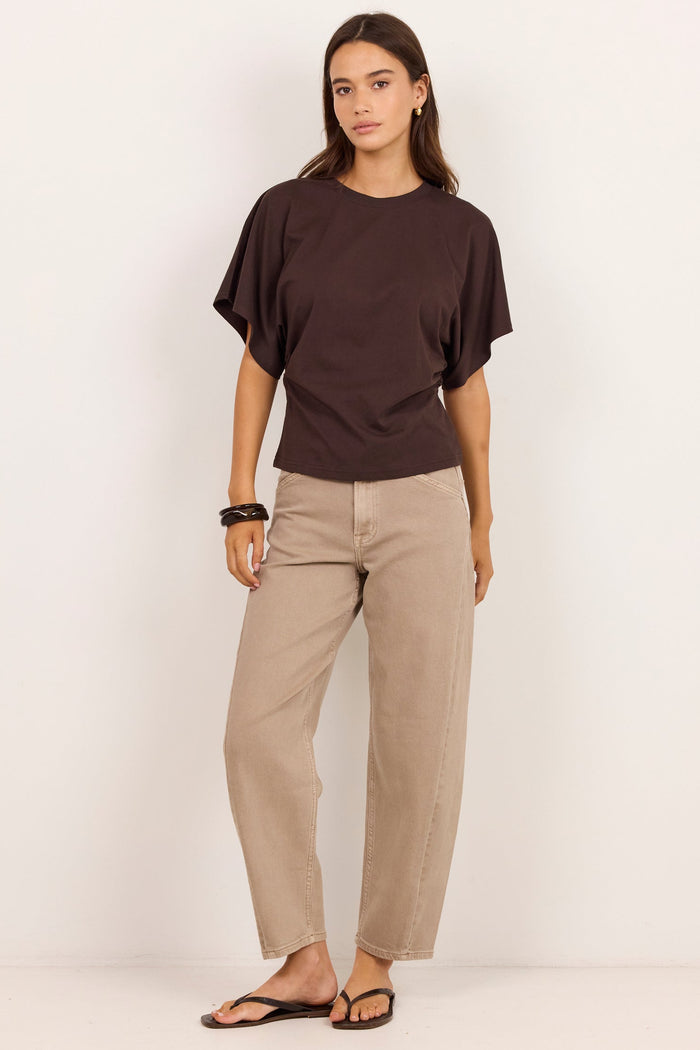 A woman modeling a dark brown short-sleeve t-shirt and beige straight-leg pants against a clean white studio background.