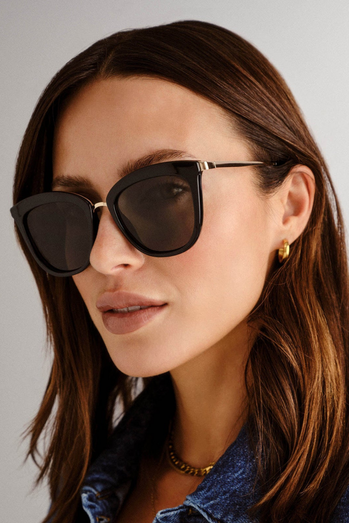 model wears cat-eye sunglasses featuring a sleek black frame paired with khaki-tinted lenses. A slim gold-tone bridge and matching gold arm accents elevate the look with a modern, minimalist touch.