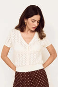 A woman wearing a cream short-sleeve knit top with a V-neck and brown wide-leg pants with white polka dots, posing against a plain off-white background.