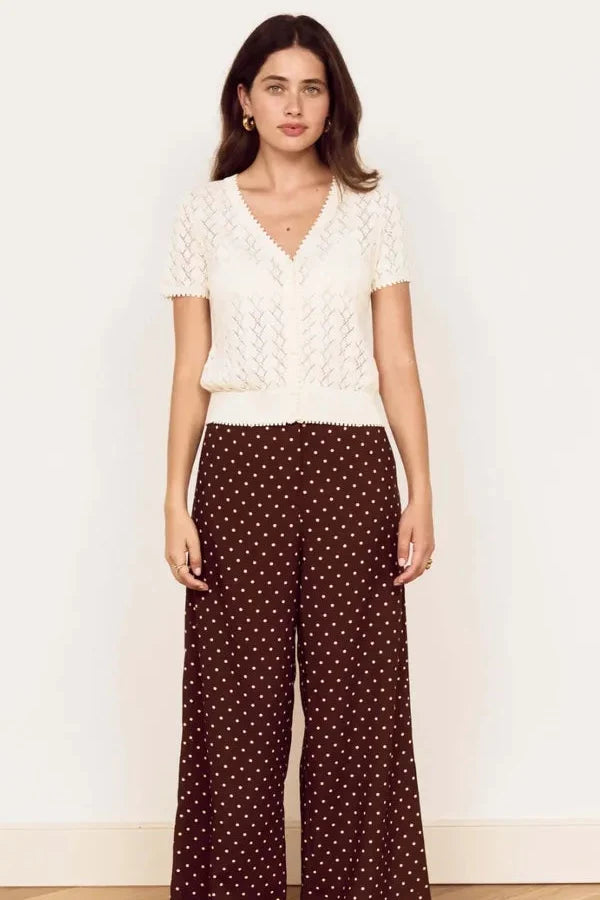 A woman stands in a studio wearing a cream short-sleeve knit top and dark brown wide-leg pants with small white polka dots, paired with black sandals.