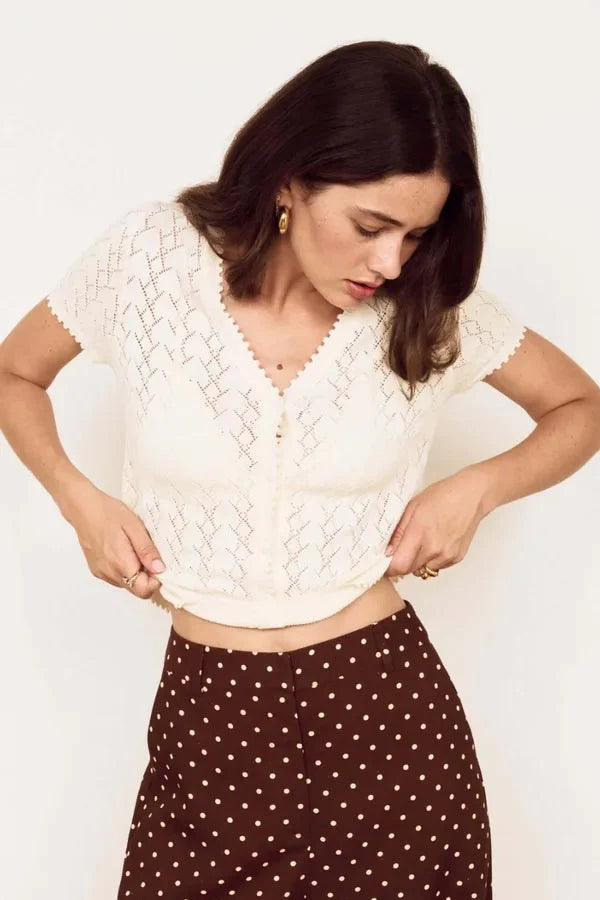 A woman wearing a short-sleeve cream lace knit top and brown pants with white polka dots, posing against a plain off-white background.